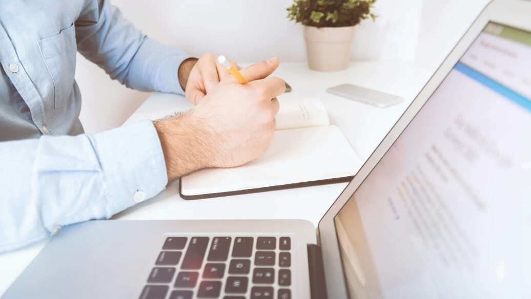 Ordinateur portable ouvert, personne écrivant dans un carnet, plante en arrière-plan.